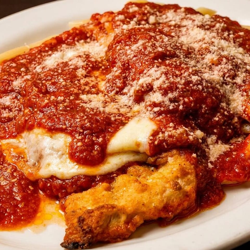 Breaded chicken parmigiana topped with melted cheese and marinara sauce, served with penne pasta on a white plate.