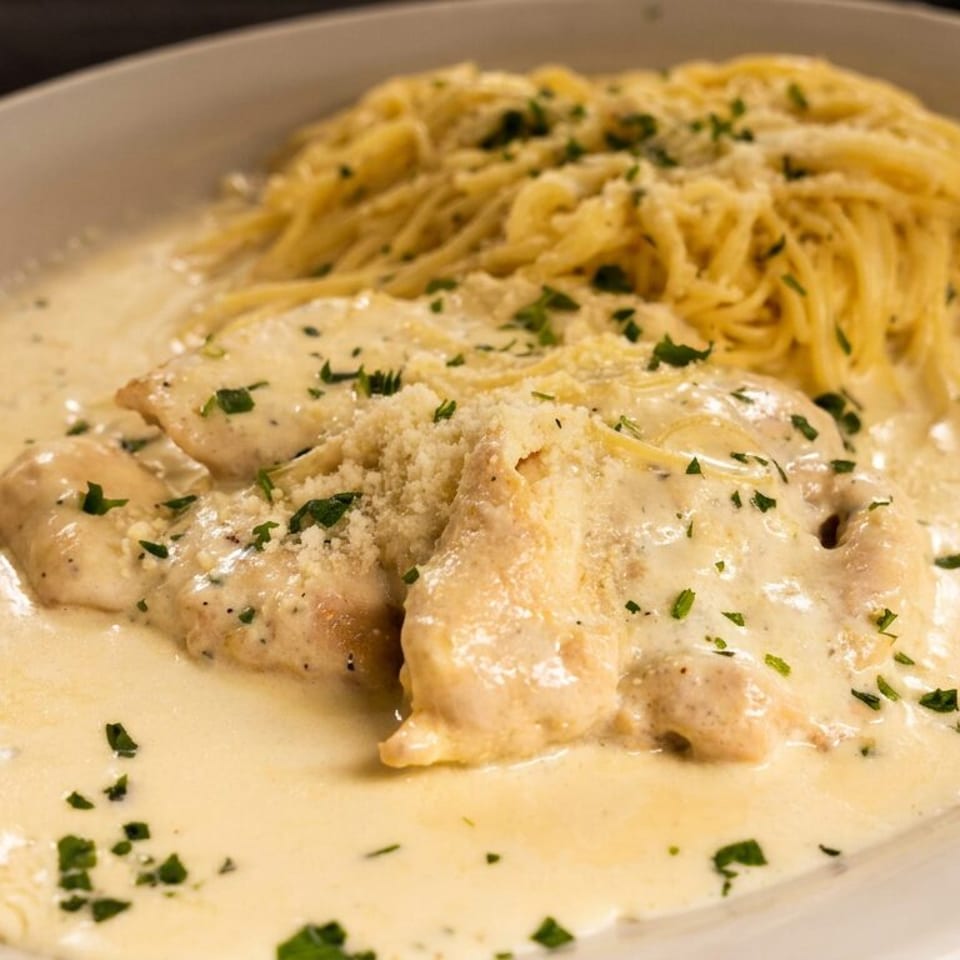Chicken breasts and capellini pasta covered in creamy sauce, garnished with parsley and grated Parmesan on a white plate.